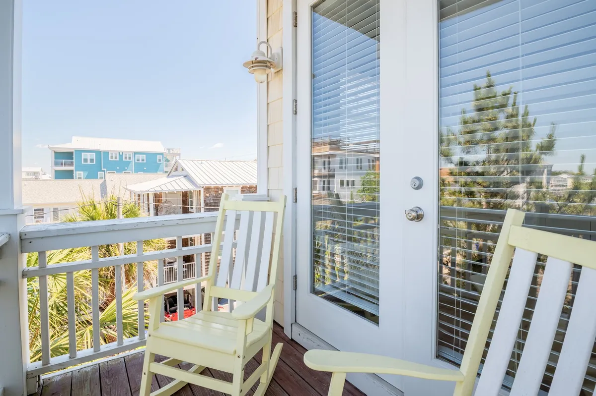 This image showcases a group of bright yellow rocking chairs on a deck, facing out towards palm trees and a vibrant blue building in the background. The glass door provides a connection between indoor and outdoor spaces, suggesting easy access for social gatherings and relaxation while enjoying the ocean breeze.