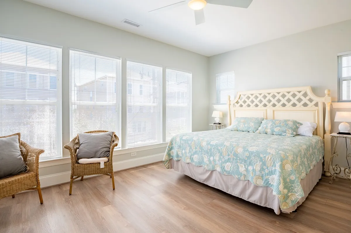 A serene bedroom with a large bed covered by a patterned quilt, two inviting wicker chairs beside expansive windows, and sunlight streaming in. The atmosphere is peaceful, cozy, and ideal for private relaxation or reading.