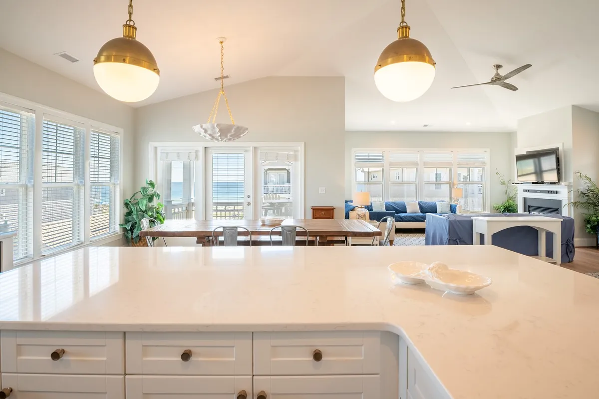 An open-concept space that seamlessly combines a kitchen, a large dining table, and a comfortable seating area. Massive windows flood the room with light, and the central kitchen island offers extra seating. The space is styled to be both inviting and functional for group activities like meals, cooking, and socializing.