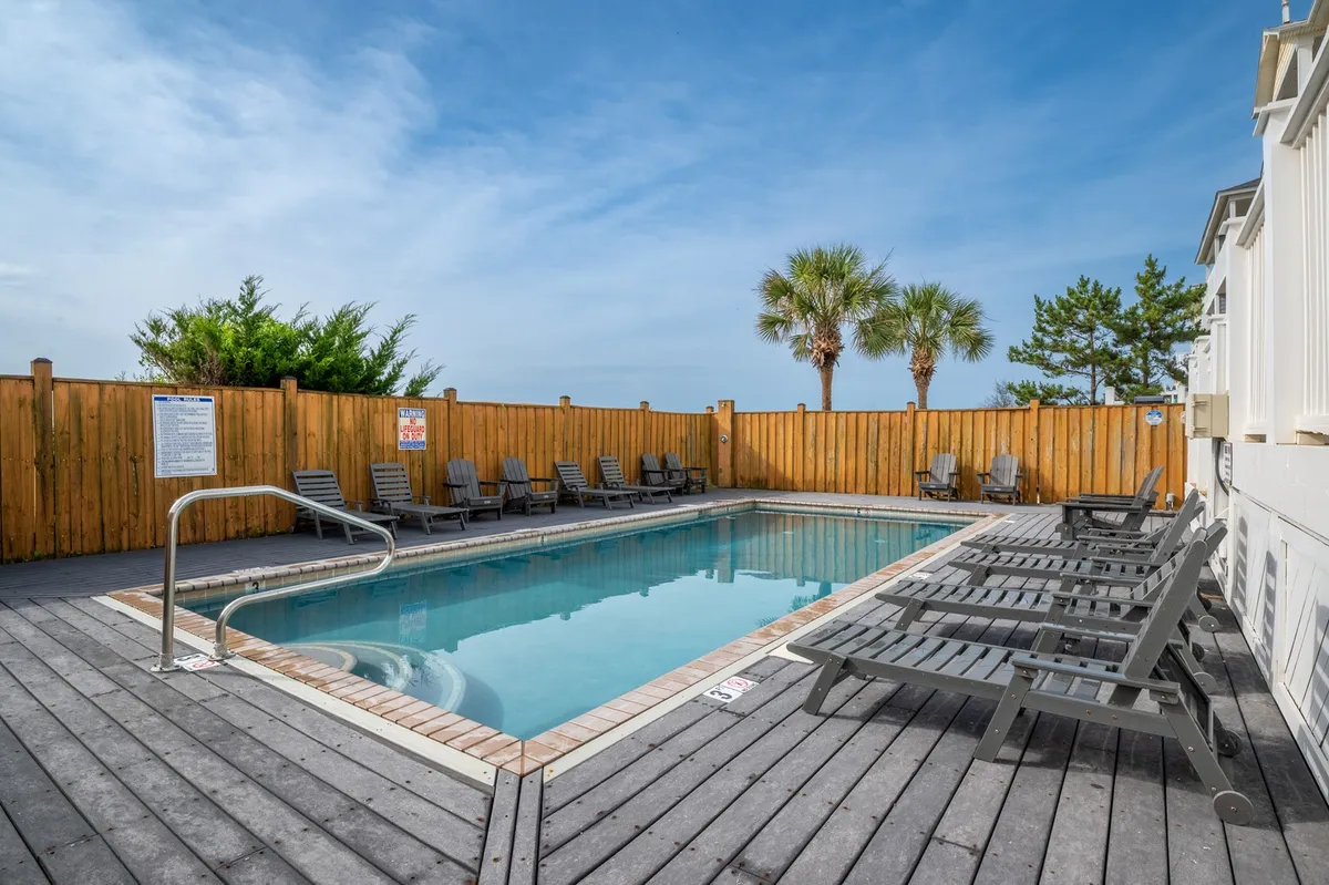 A sparkling community swimming pool framed by palm trees and a wooden fence. Several lounge chairs line the sides, inviting guests to sunbathe, relax, or socialize poolside. The scene radiates leisure and comfort, encapsulating the essence of vacation relaxation.