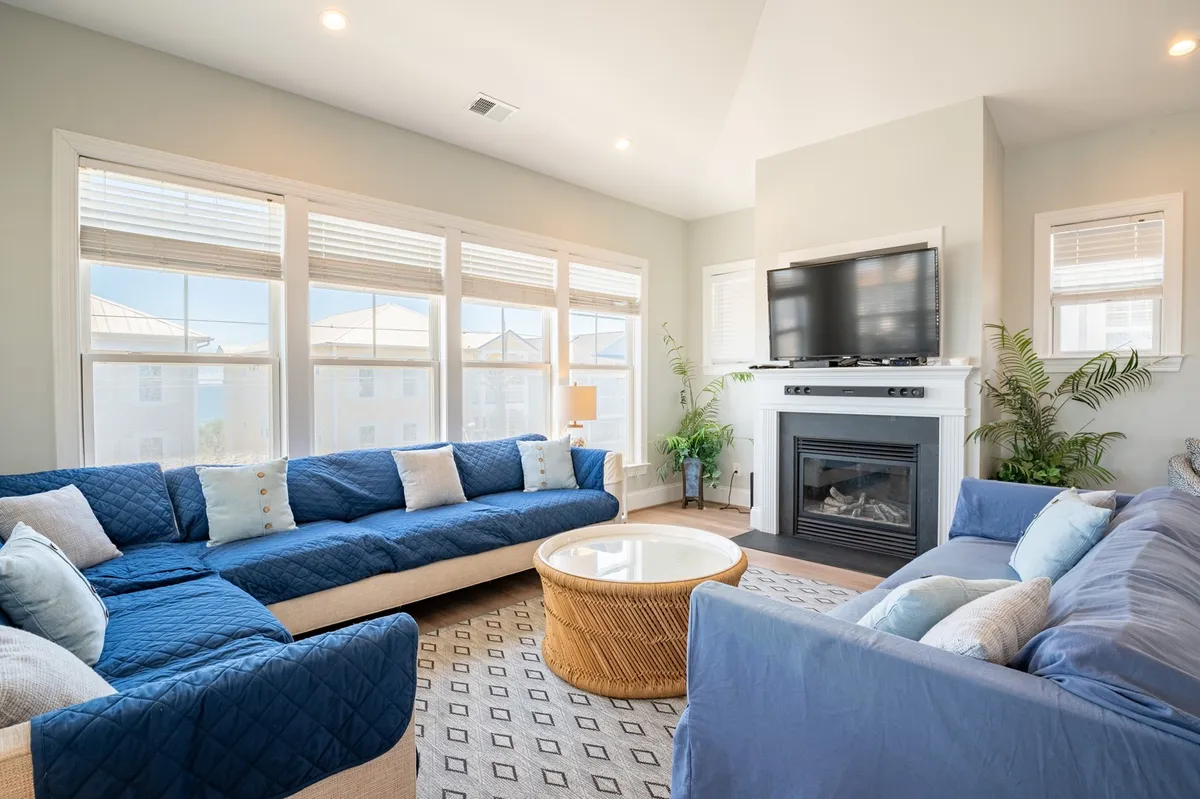 A spacious, sunlit great room featuring large sectional sofas centered around a modern gas fireplace and a coffee table. Floor-to-ceiling windows let in abundant natural light, and a TV is mounted above the fireplace. The overall mood is cozy and welcoming, perfect for family and friends to gather for movie nights or relaxed conversations.
