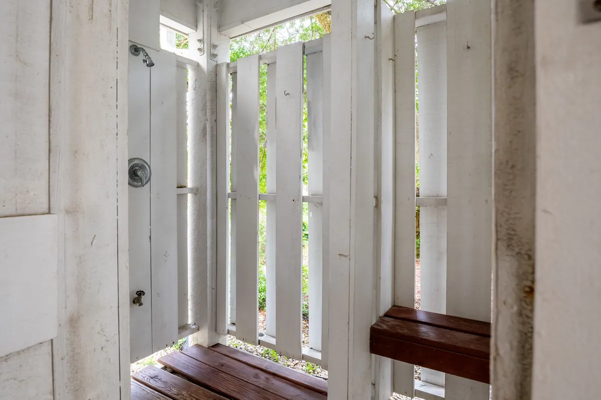 A refreshing outdoor shower with a wooden bench and open shutters, surrounded by lush greenery. The setup provides privacy while maintaining a connection to the outdoors—perfect for rinsing off after the beach.