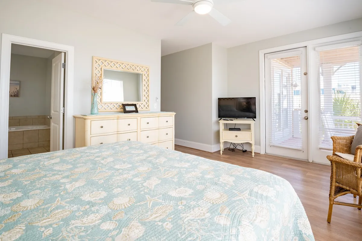 A spacious, light-filled bedroom featuring a comfortable bed, dresser, and mirror, with glass balcony doors opening to an inviting outdoor space. Subtle décor and a calm palette create a restful atmosphere, while the doors suggest deck access and potential for water views.