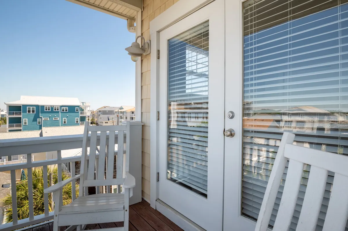 This image features white rocking chairs placed on a porch or balcony, with a glass door leading indoors and a view of neighboring houses visible beyond the railing. The scene is serene and welcoming, hinting at the enjoyment of sunsets, morning coffee, and relaxed conversations while overlooking the neighborhood.