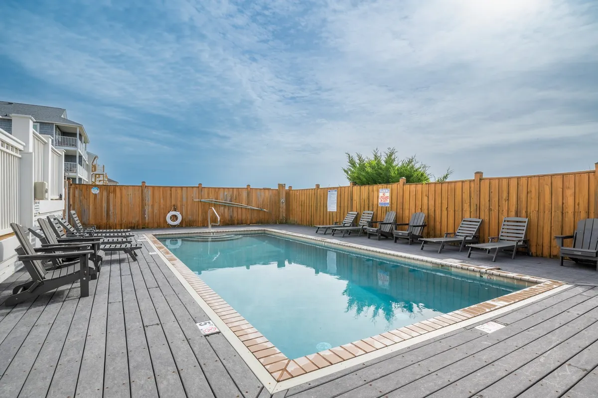 A tranquil swimming pool area featuring lounge chairs and a wooden fence, ideal for sunbathing, swimming, and relaxing after a day at the beach. The pool area feels welcoming and safe for families.