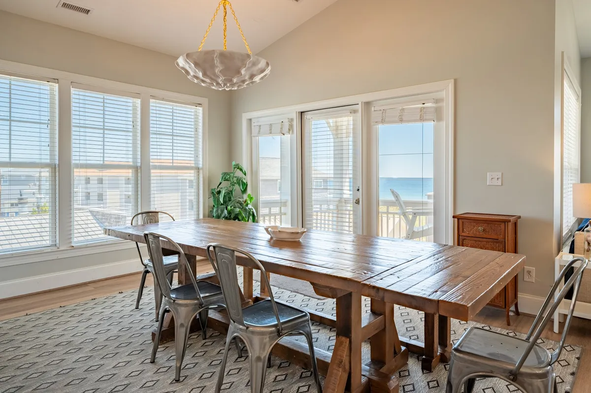 A warm, welcoming dining area with a large wooden farm table, surrounded by chairs and illuminated by a chandelier. Floor-to-ceiling windows offer a stunning ocean view, reinforcing the sense of openness and togetherness. The space is perfect for both casual group breakfasts and festive dinners.