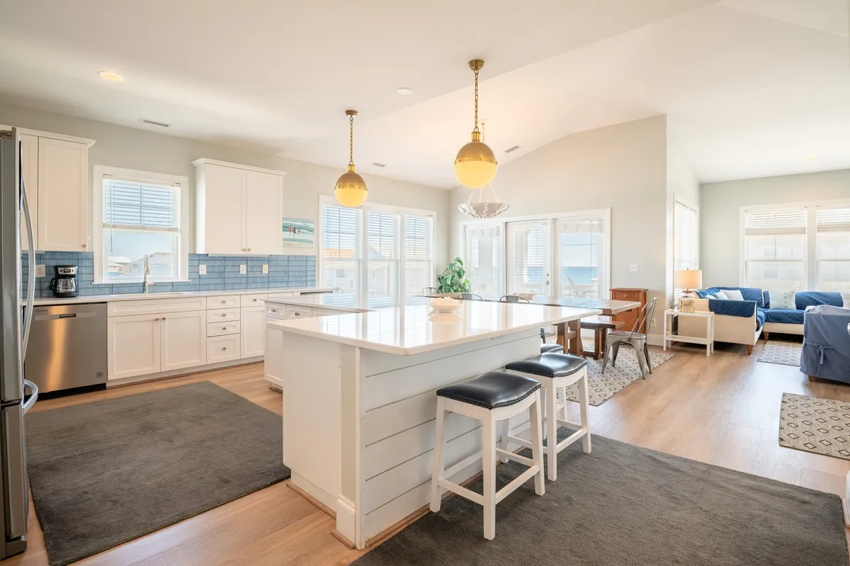 A spacious, open-plan living and kitchen area is shown, with a large kitchen island, blue backsplash, and comfortable living space in view. Large windows provide natural light, while decorative pendant lights and bar seating suggest a place for group meals, cooking, and relaxation.